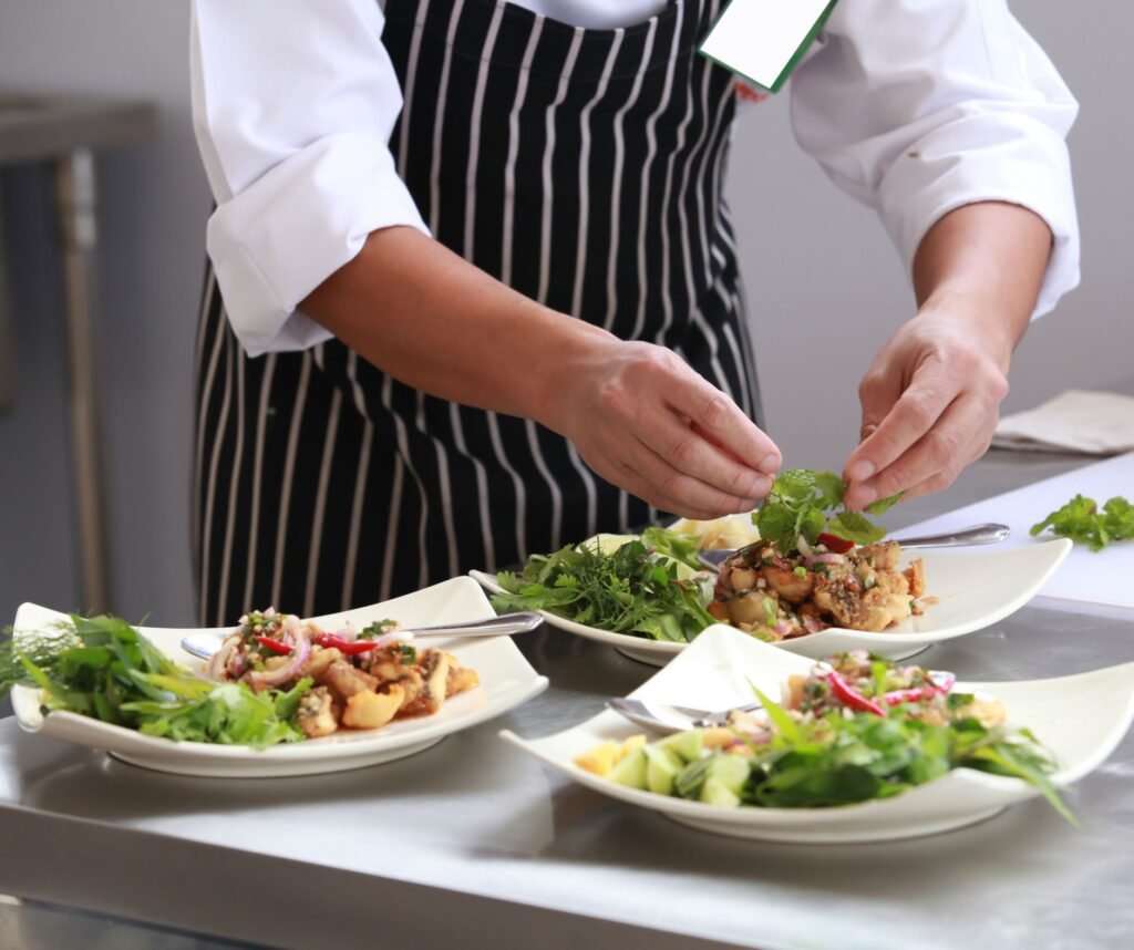 Professional chef in a striped apron carefully garnishing three gourmet seafood salad plates with fresh herbs.