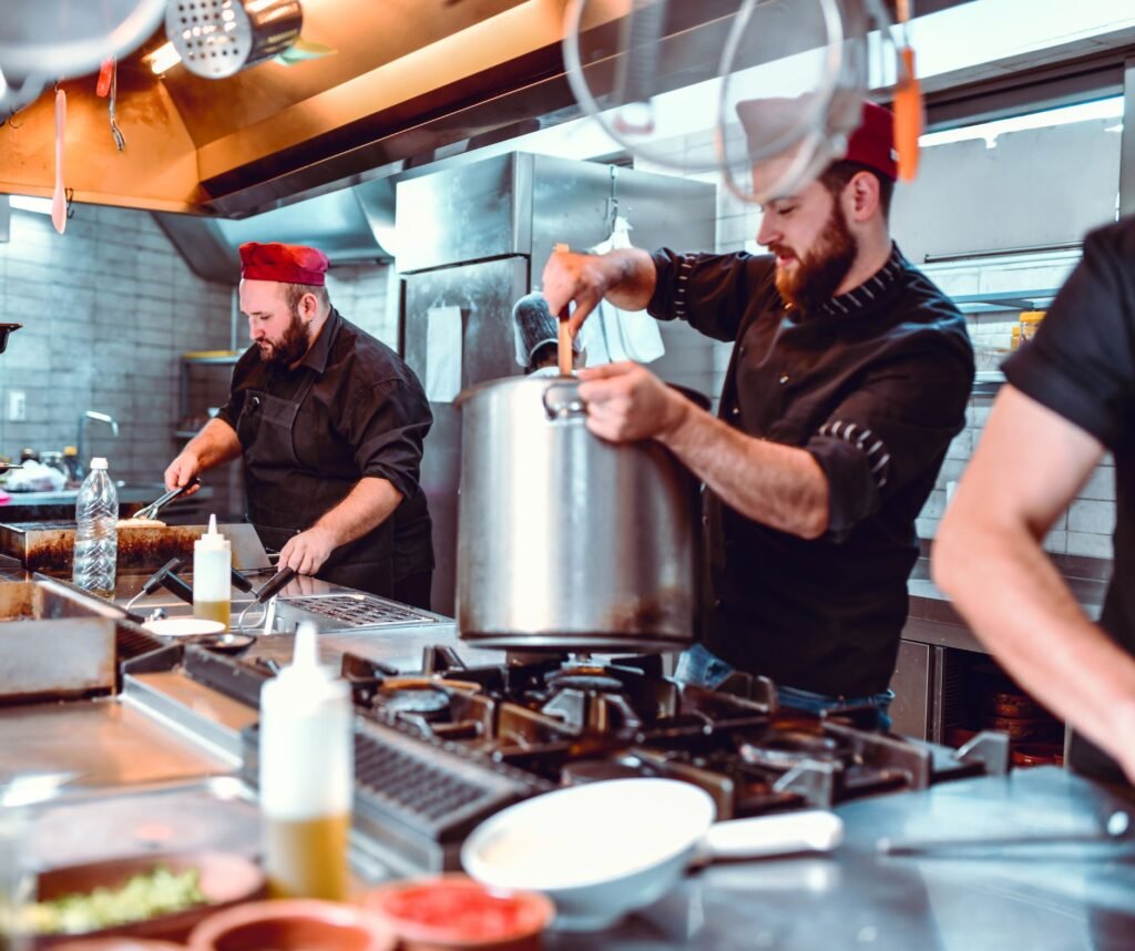 A restaurant kitchen team preparing food with stainless steel appliances, industrial stovetops, and various cooking ingredients in the foreground.
