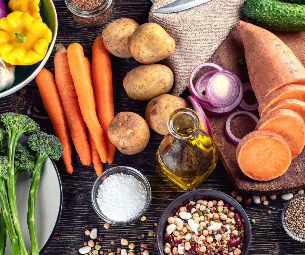 Overhead view of fresh vegetables and pantry ingredients on a wooden table, including carrots, potatoes, sliced sweet potato, broccoli, red onion, and olive oil.